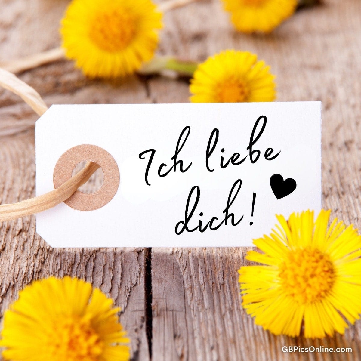 Weiße Geschenkanhänger mit Schrift „Ich liebe dich!“ und Herz, umgeben von gelben Löwenzahnblüten auf Holz