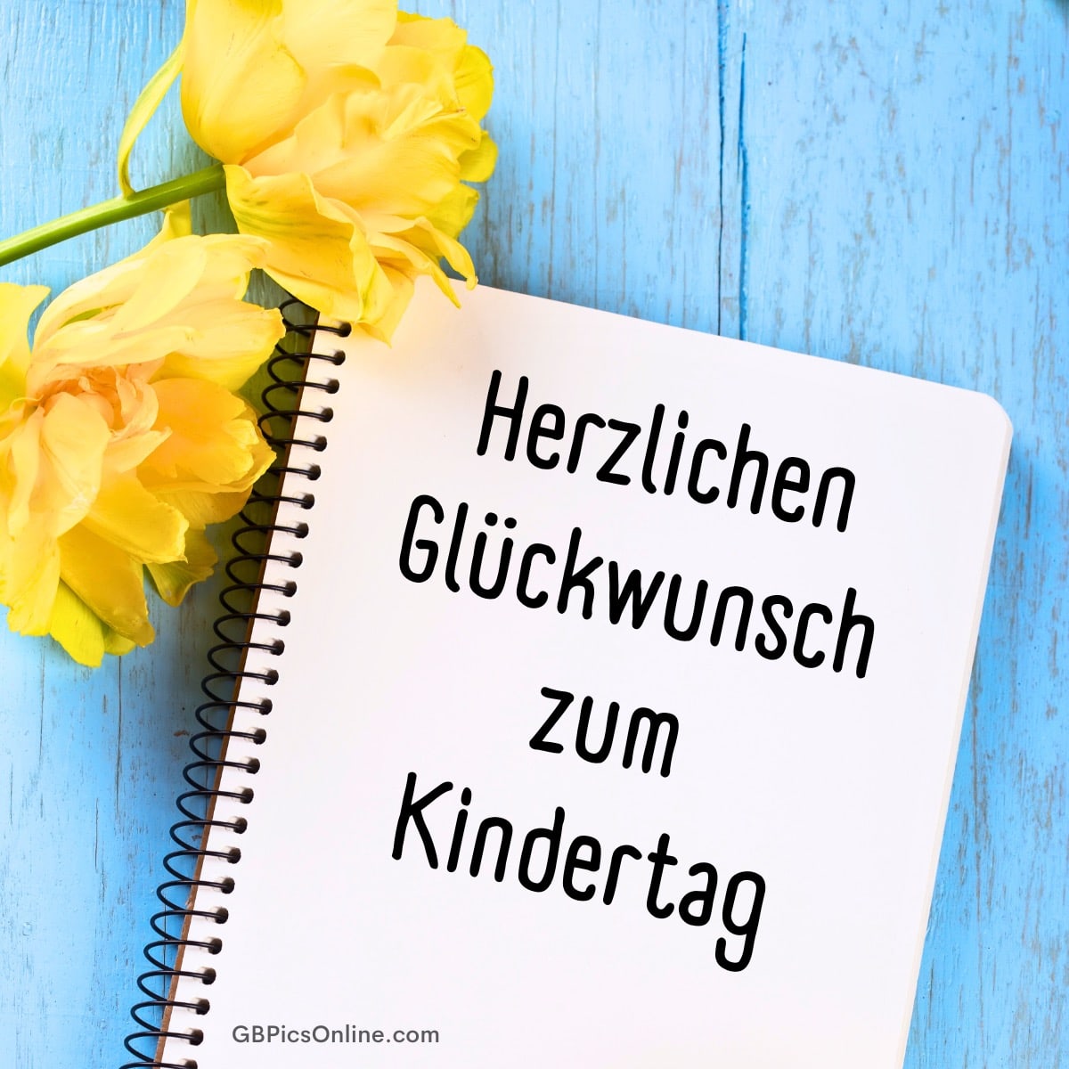 Zwei gelbe Blumen neben einem Spiralblock mit der Aufschrift „Herzlichen Glückwunsch zum Kindertag“ auf blauem Holz