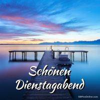 Steg führt in einen stillen See bei Sonnenuntergang; bunter Himmel, Berge am Horizont, Schriftzug „Schönen Dienstagabend“