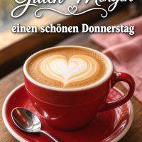 Rote Tasse Cappuccino mit Herz-Latte-Art auf Holz­tisch, dazu Löffel und Morgengruß
