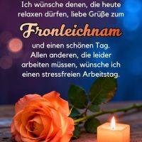 Grußtext zu Fronleichnam über einer orangefarbenen Rose und einer brennenden Kerze auf einem Holztisch