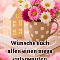 Bunter Blumenstrauß in gepunkteter Tasse neben kleinem Dekohaus auf hellem Tablett vor rosa Hintergrund